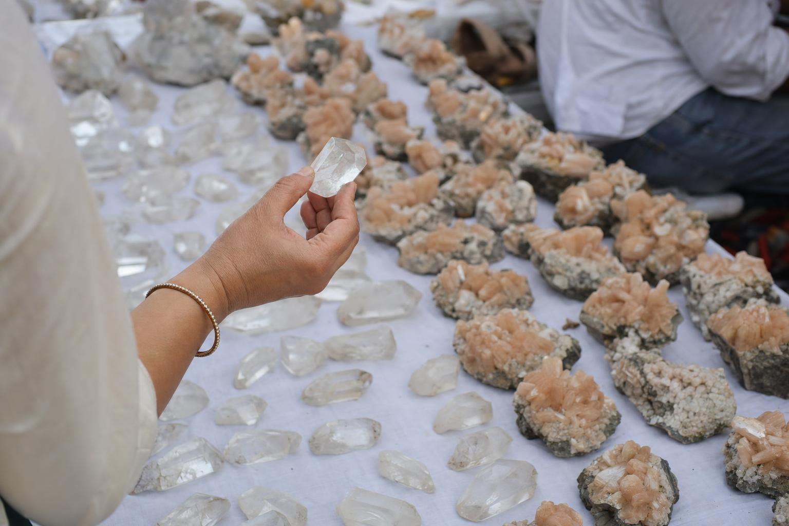 A hand lifts a clear crystal to the window light above rows of raw crystals on white cloth.